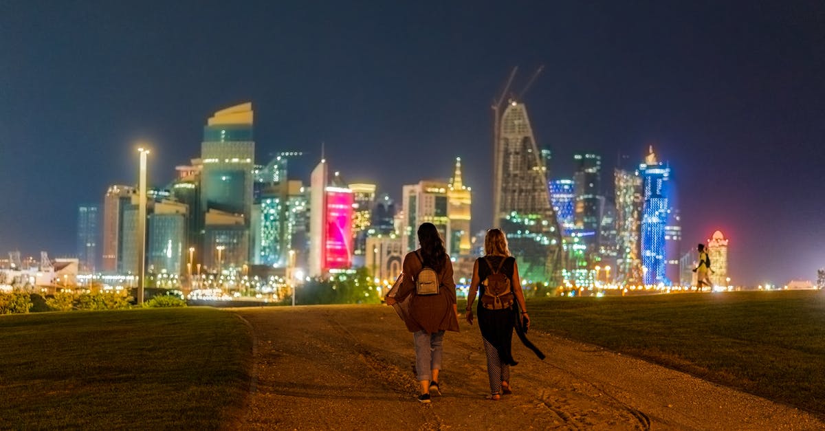 Qatar - Doha - Lounge Options - Back view of unrecognizable female tourists in casual clothes and backpack walking on sandy road towards modern illuminated buildings in Doha at night Qatar - Doha - Lounge Options - Back view of unrecognizable female tourists in casual clothes and backpack walking on sandy road towards modern illuminated buildings in Doha at night