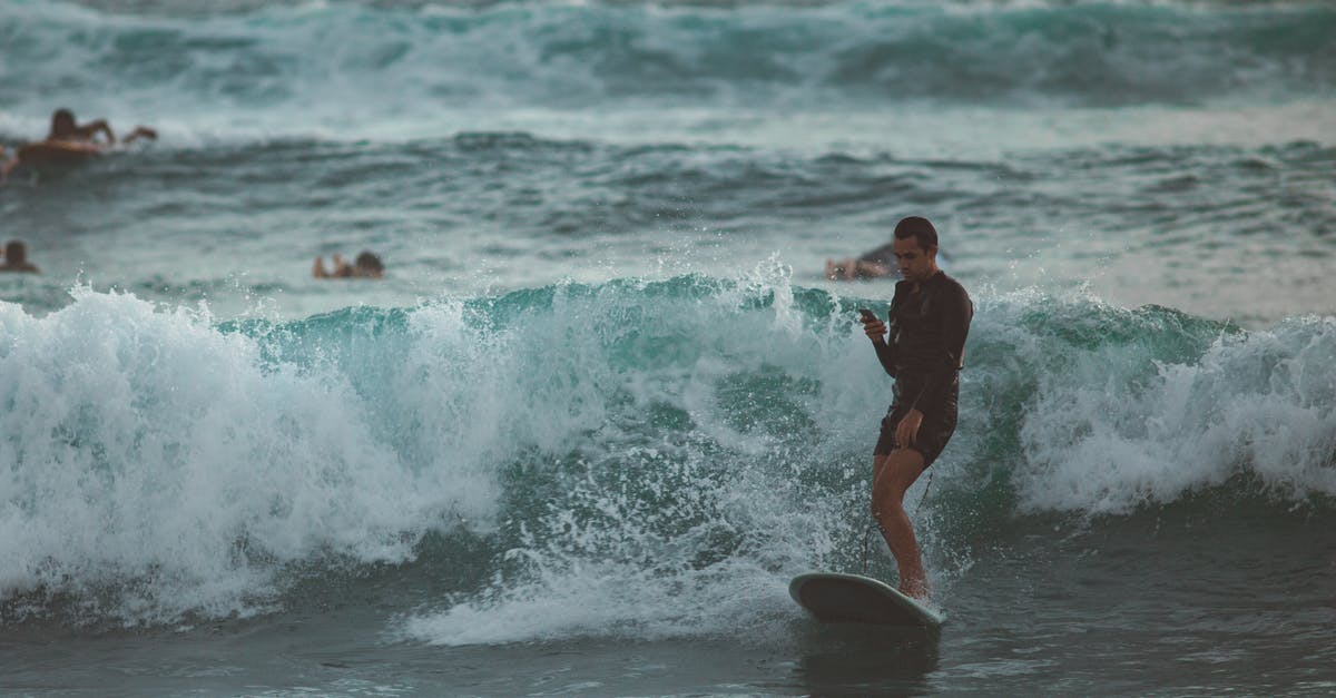 Re-entry to Germany after vacation using blue card - Young male surfer in short wetsuit riding surfboard and using mobile in magnificent restless sea with other surfers paddling behind Re-entry to Germany after vacation using blue card - Young male surfer in short wetsuit riding surfboard and using mobile in magnificent restless sea with other surfers paddling behind