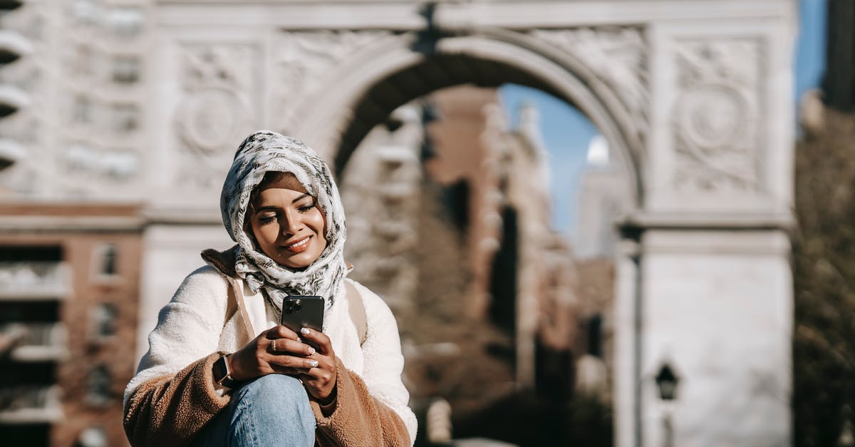 Re-entry to Germany after vacation using blue card - Self assured young ethnic female in stylish clothes and Muslim headscarf smiling while messaging on smartphone sitting in city square near aged arch Re-entry to Germany after vacation using blue card - Self assured young ethnic female in stylish clothes and Muslim headscarf smiling while messaging on smartphone sitting in city square near aged arch