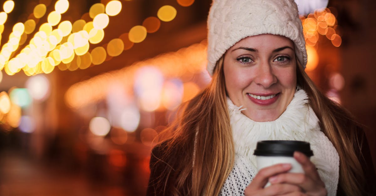 Rent clothing in Frankfurt / Being warm without cold weather clothing - Cheerful female in warm outerwear standing with beverage to go on street and chilling during weekend in evening in city illumination while looking at camera Rent clothing in Frankfurt / Being warm without cold weather clothing - Cheerful female in warm outerwear standing with beverage to go on street and chilling during weekend in evening in city illumination while looking at camera