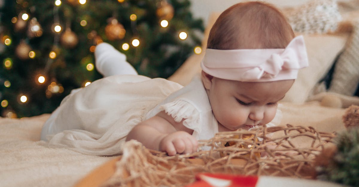 Renting a holiday house in the Riviera with a small child - Toddler girl in white dress lying on comfortable bed near pillows and decorative objects in light bedroom with Christmas tree with baubles and garland at home Renting a holiday house in the Riviera with a small child - Toddler girl in white dress lying on comfortable bed near pillows and decorative objects in light bedroom with Christmas tree with baubles and garland at home