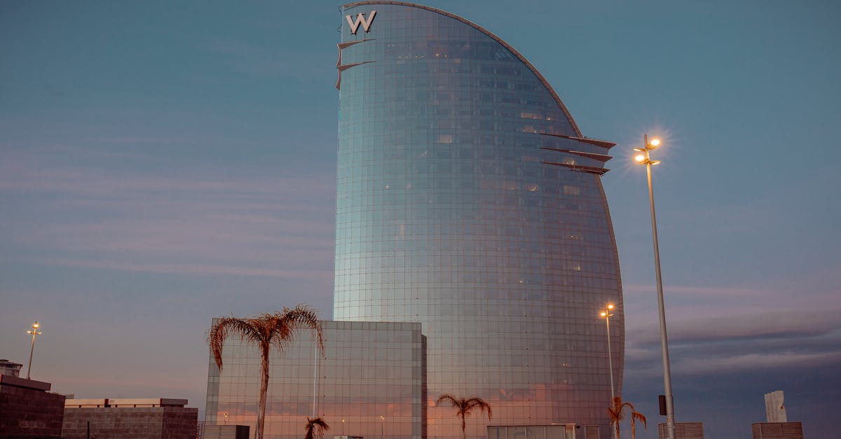 Reserve hotel from Barcelona airport? - Contemporary building facade in form of sail against streetlights and anonymous people walking under sunset sky in Barcelona Spain Reserve hotel from Barcelona airport? - Contemporary building facade in form of sail against streetlights and anonymous people walking under sunset sky in Barcelona Spain