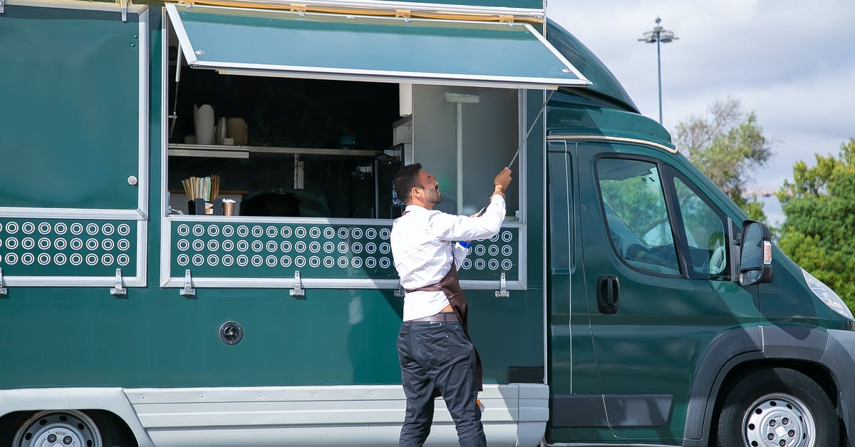 Roland-Garros (French Open) public viewing in Paris? - Side view young waiter in apron standing outside food truck and opening counter door in green lush park Roland-Garros (French Open) public viewing in Paris? - Side view young waiter in apron standing outside food truck and opening counter door in green lush park