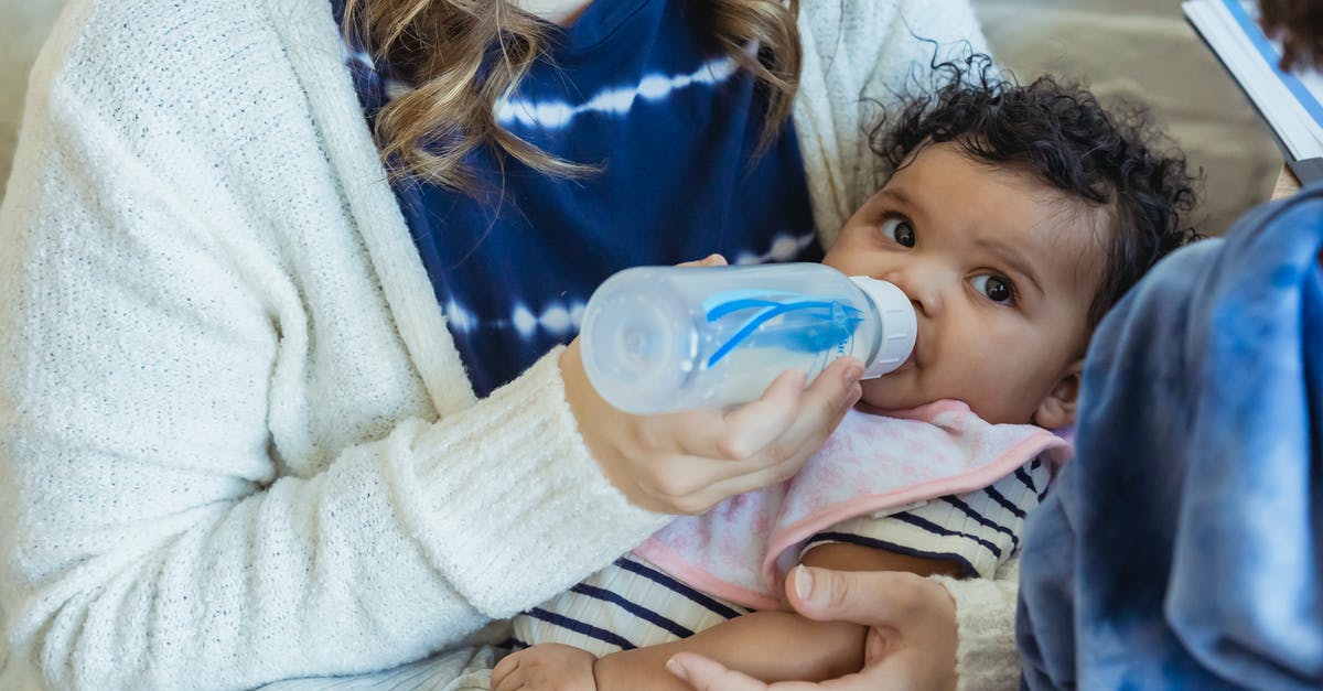 Russian mom traveling from US to Russian Federation with little kid [closed] - High angle of crop mother embracing little African American baby and feeding from bottle sitting on sofa near son