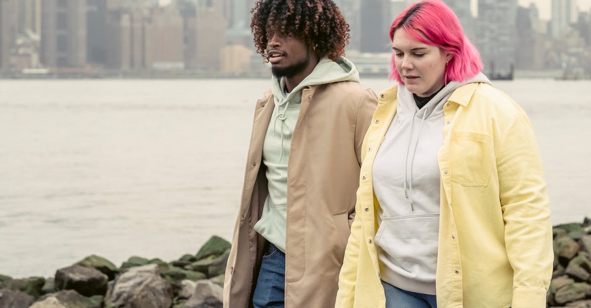 Send 1 overweight suitcase instead of 2 underweight? - Multiracial couple in stylish clothes strolling on coast with stones near calm river against residential buildings during date in city Send 1 overweight suitcase instead of 2 underweight? - Multiracial couple in stylish clothes strolling on coast with stones near calm river against residential buildings during date in city