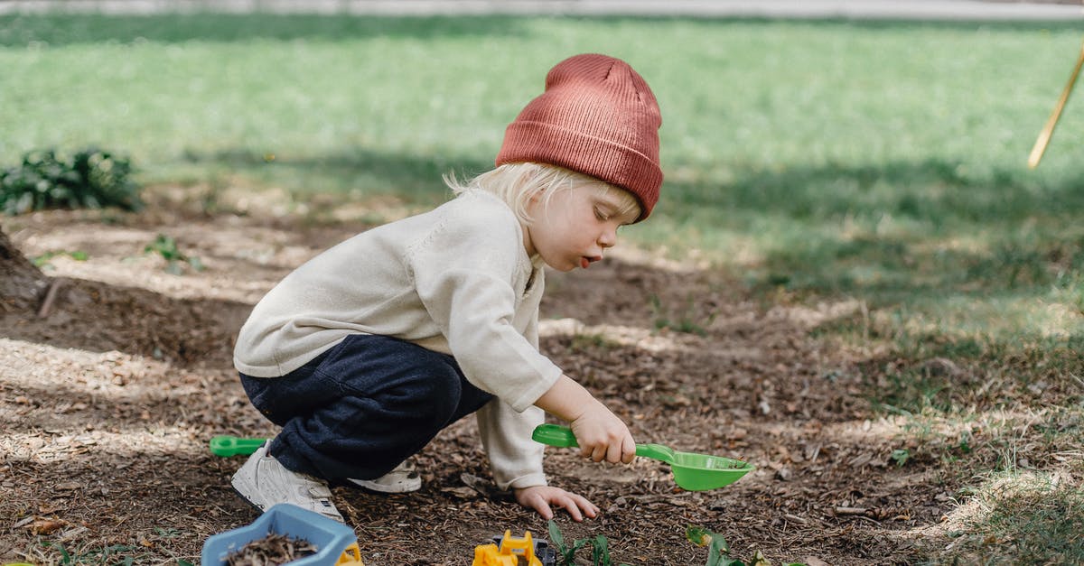 Small layover time. What to do? - Side view of little boy in casual clothes and brown hat playing with plastic toys in backyard Small layover time. What to do? - Side view of little boy in casual clothes and brown hat playing with plastic toys in backyard