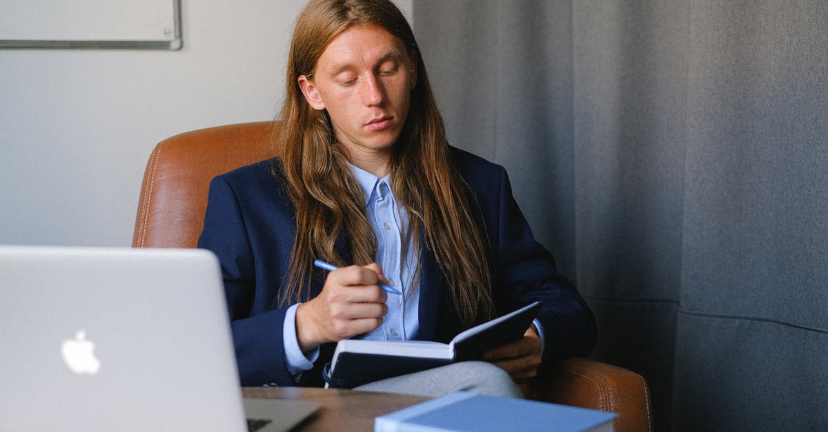 Strategy for cruise tickets: book now or last minute? - Pensive androgynous businessman in formal suit sitting at table with laptop and book and taking notes in notebook in workspace Strategy for cruise tickets: book now or last minute? - Pensive androgynous businessman in formal suit sitting at table with laptop and book and taking notes in notebook in workspace