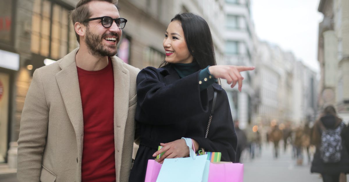 Tax Free shopping in Sweden departing from Copenhagen with a stopover in Helsinki - From below happy stylish Asian woman in warm clothes smiling at cheerful unshaven man in glasses and pointing finger away while walking along street together in city after shopping Tax Free shopping in Sweden departing from Copenhagen with a stopover in Helsinki - From below happy stylish Asian woman in warm clothes smiling at cheerful unshaven man in glasses and pointing finger away while walking along street together in city after shopping