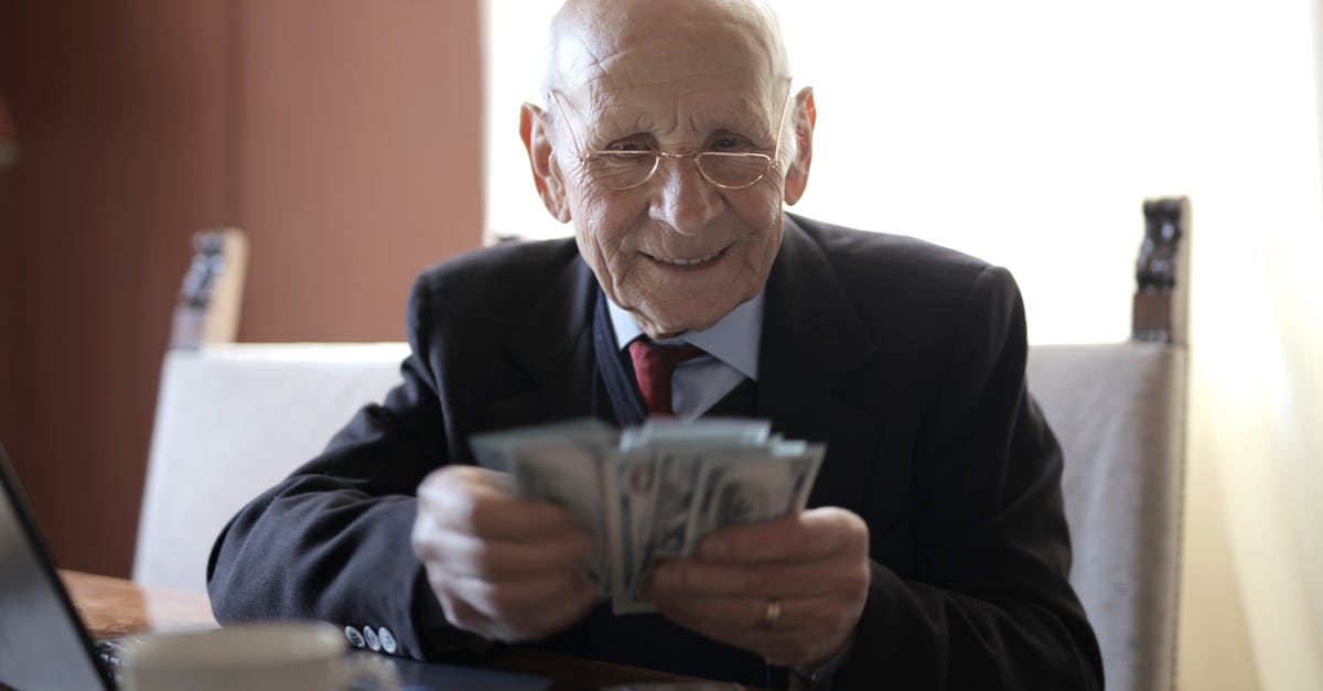 Toll booth bill by mail - Positive senior businessman in formal suit and eyeglasses counting money bills while sitting at wooden table with cup of beverage and near opened laptop Toll booth bill by mail - Positive senior businessman in formal suit and eyeglasses counting money bills while sitting at wooden table with cup of beverage and near opened laptop