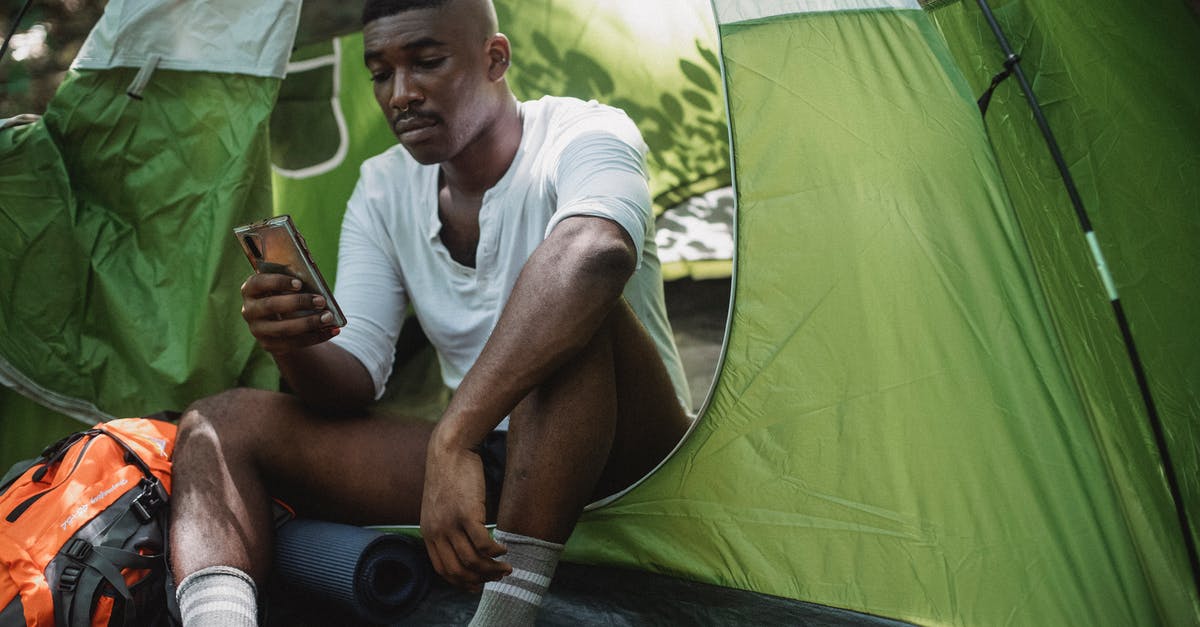 Tourist cell phone use in Myanmar - Sad African American male traveler resting in tent and browsing Internet on cellphone while spending journey in nature Tourist cell phone use in Myanmar - Sad African American male traveler resting in tent and browsing Internet on cellphone while spending journey in nature