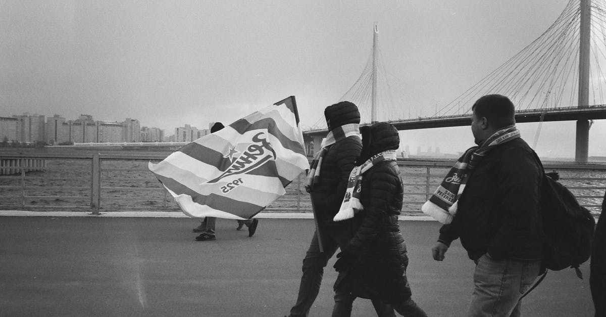 Tourist Schengen visa dates do not match itinerary - Black and white side view of people in warm clothes with flag walking along river embankment against modern bridge Tourist Schengen visa dates do not match itinerary - Black and white side view of people in warm clothes with flag walking along river embankment against modern bridge