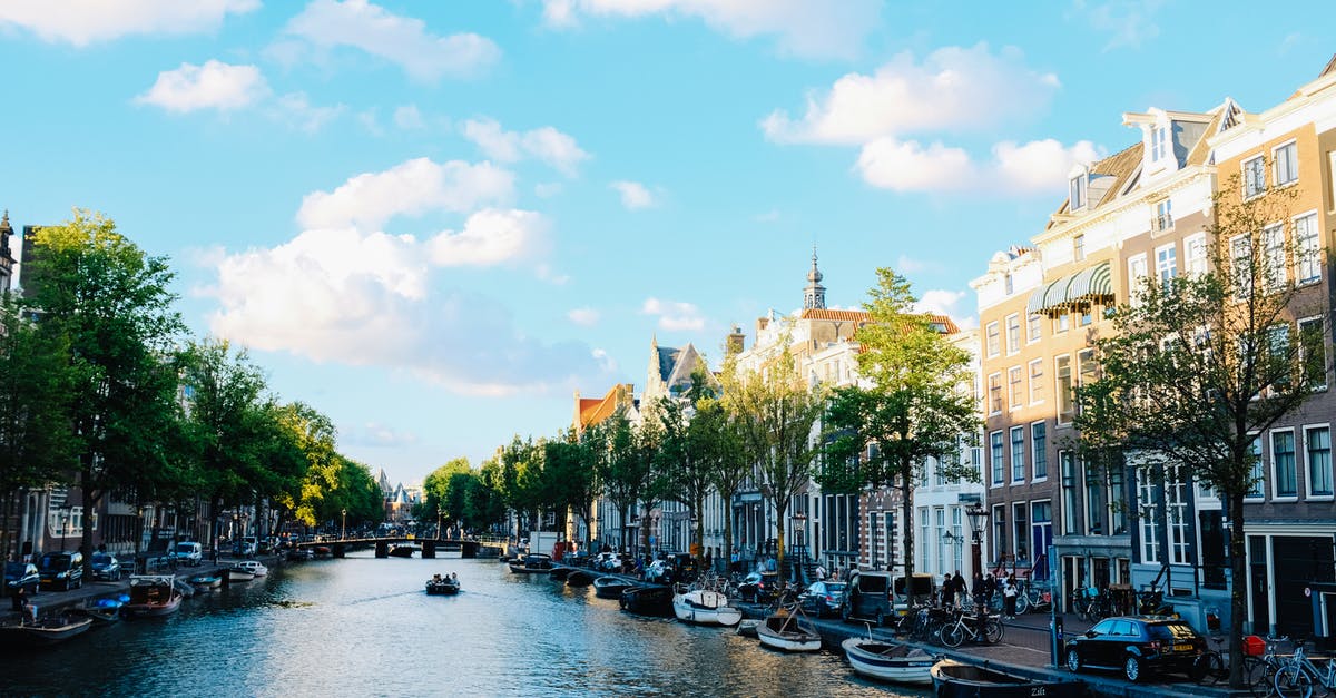 Transportation in Netherlands: to and around Amsterdam - Magnificent cityscape of Amsterdam with traditional boats moored on calm river near historic buildings under cloudy blue sky Transportation in Netherlands: to and around Amsterdam - Magnificent cityscape of Amsterdam with traditional boats moored on calm river near historic buildings under cloudy blue sky