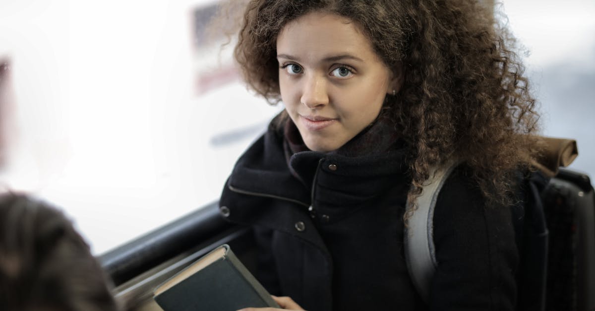 Travelling to Oman by Bus from Dubai - From above view of positive young lady with curly hair in casual warm coat sitting with book on passenger seat near window in bus and looking at camera Travelling to Oman by Bus from Dubai - From above view of positive young lady with curly hair in casual warm coat sitting with book on passenger seat near window in bus and looking at camera