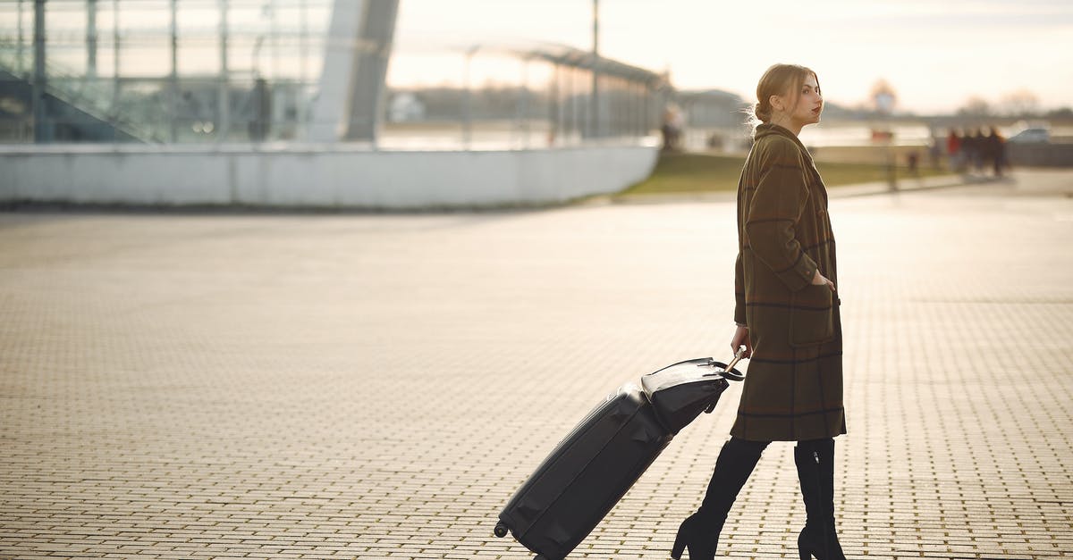 Two bag limit on Hand/Carry On Baggage at Heathrow? - Side view of full length young female traveler in trendy plaid brown coat and high boots with suitcase walking on pavement near modern building on city street in morning Two bag limit on Hand/Carry On Baggage at Heathrow? - Side view of full length young female traveler in trendy plaid brown coat and high boots with suitcase walking on pavement near modern building on city street in morning
