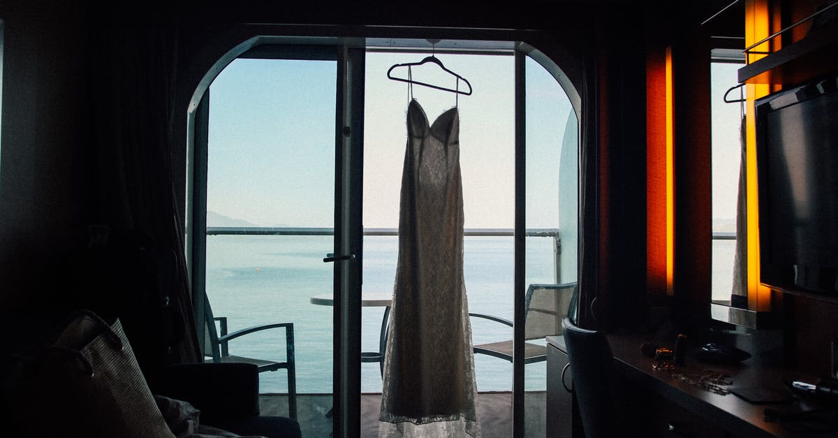UK Visit Visa hotel arrangement - Interior of resort room in darkness with white dress on hanger on window against ocean UK Visit Visa hotel arrangement - Interior of resort room in darkness with white dress on hanger on window against ocean