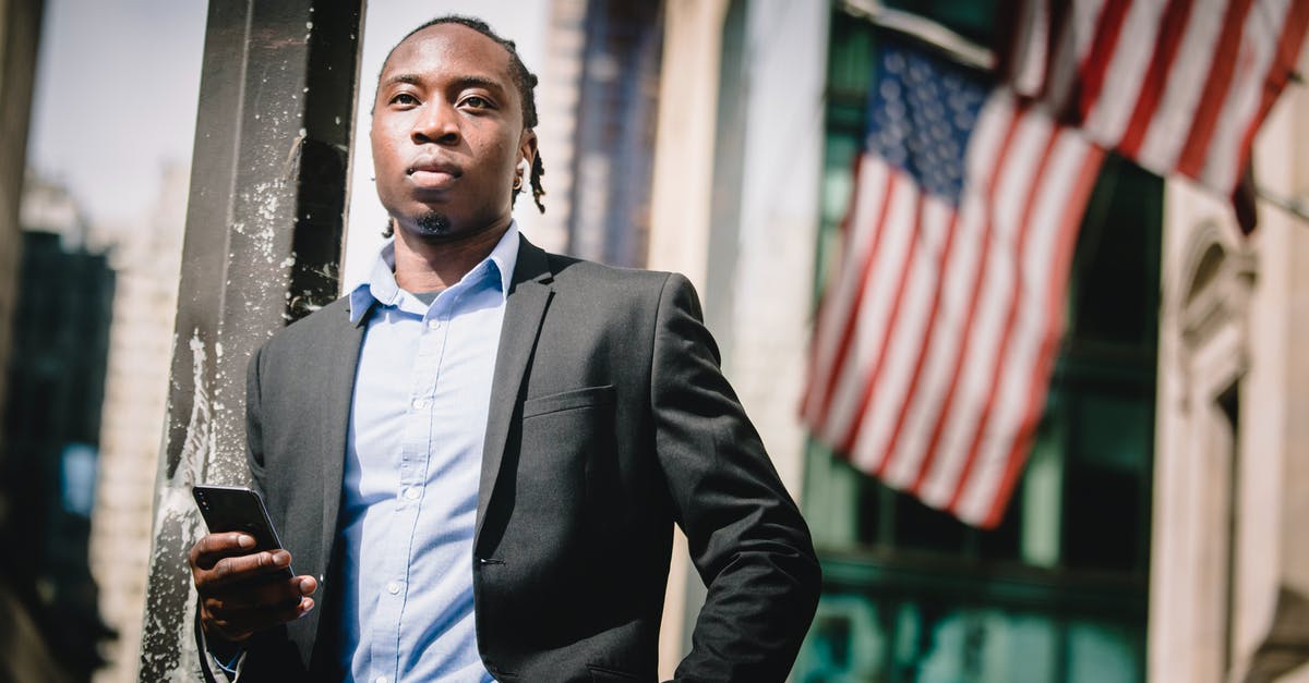 US non-immigrant visa appointment at Frankfurt - Low angle of serious well dressed ethnic male manager leaning on metal pillar on street and using cellphone against government building with hanging American flags