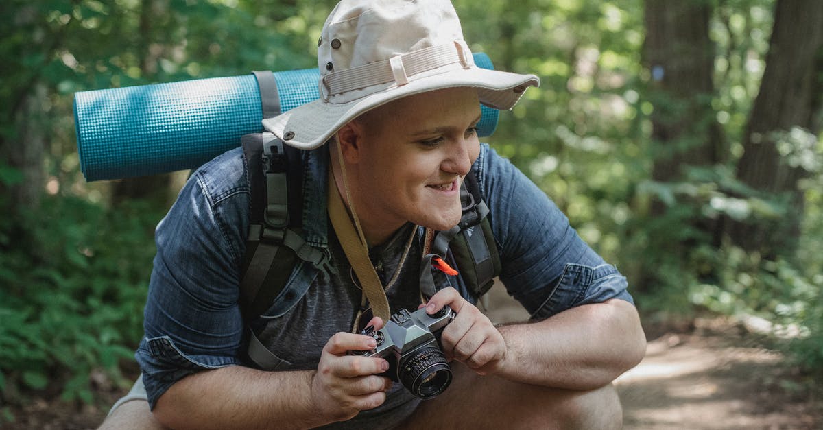 Using squat toilets with arthritic knees - Young smiling male hiker with mat and photo camera squatting on pathway with trees behind while looking away in woods Using squat toilets with arthritic knees - Young smiling male hiker with mat and photo camera squatting on pathway with trees behind while looking away in woods
