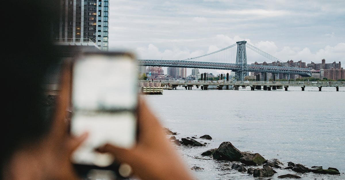What coast or river-adjacent city is in this photo? - Crop anonymous African American male standing on embankment and taking picture of skyscrapers and bridge What coast or river-adjacent city is in this photo? - Crop anonymous African American male standing on embankment and taking picture of skyscrapers and bridge