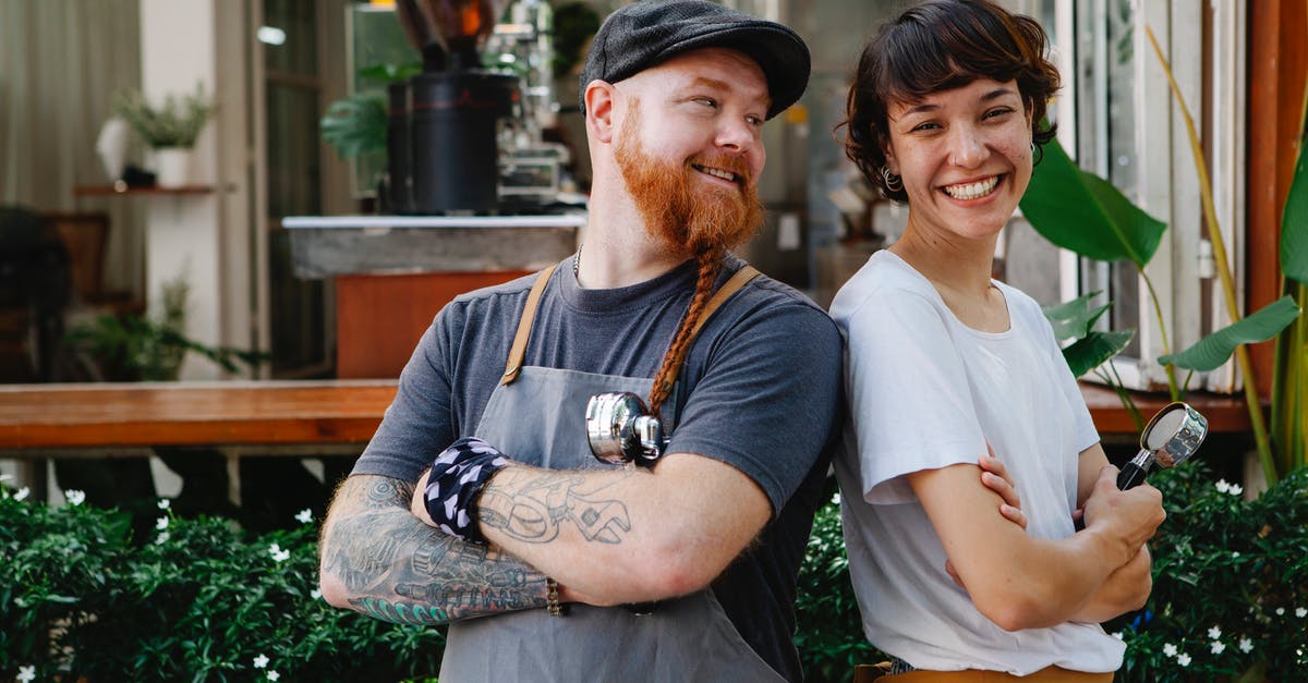 What happens if you don't register your local address when visiting Germany? - Couple of smiling barista coworkers in casual outfit and aprons standing with portafilter near cafeteria in street near plants in daylight