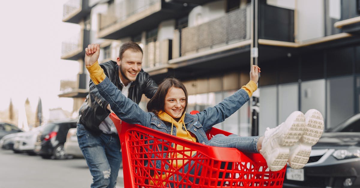 Where can I ride a pump trolley? - Cheerful adult male in casual outfit riding happy girl in jeans clothes on shopping trolley on parking on city street near modern multistory building Where can I ride a pump trolley? - Cheerful adult male in casual outfit riding happy girl in jeans clothes on shopping trolley on parking on city street near modern multistory building