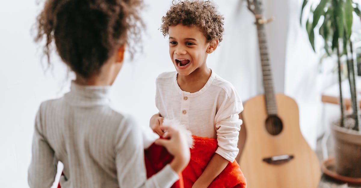 Winter apparel for Europe (November-December) - Funny African American boy looking at anonymous sister while standing with Santa Claus costumes in room with guitar during Christmas holiday Winter apparel for Europe (November-December) - Funny African American boy looking at anonymous sister while standing with Santa Claus costumes in room with guitar during Christmas holiday